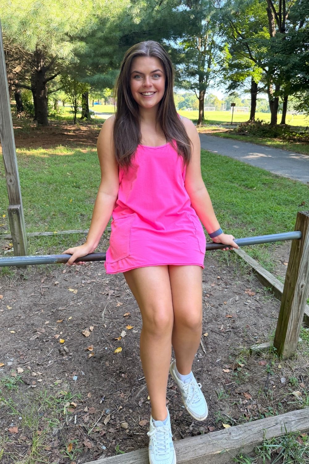 A woman wearing a bright pink sleeveless mini dress and white sneakers stands outdoors on a wooden structure in a park, holding a metal bar with her hands.