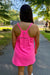 A woman viewed from behind wearing a bright pink athletic tank dress with thin straps and a small zippered pocket on the back, standing on an outdoor paved path with trees and a tennis court in the background.