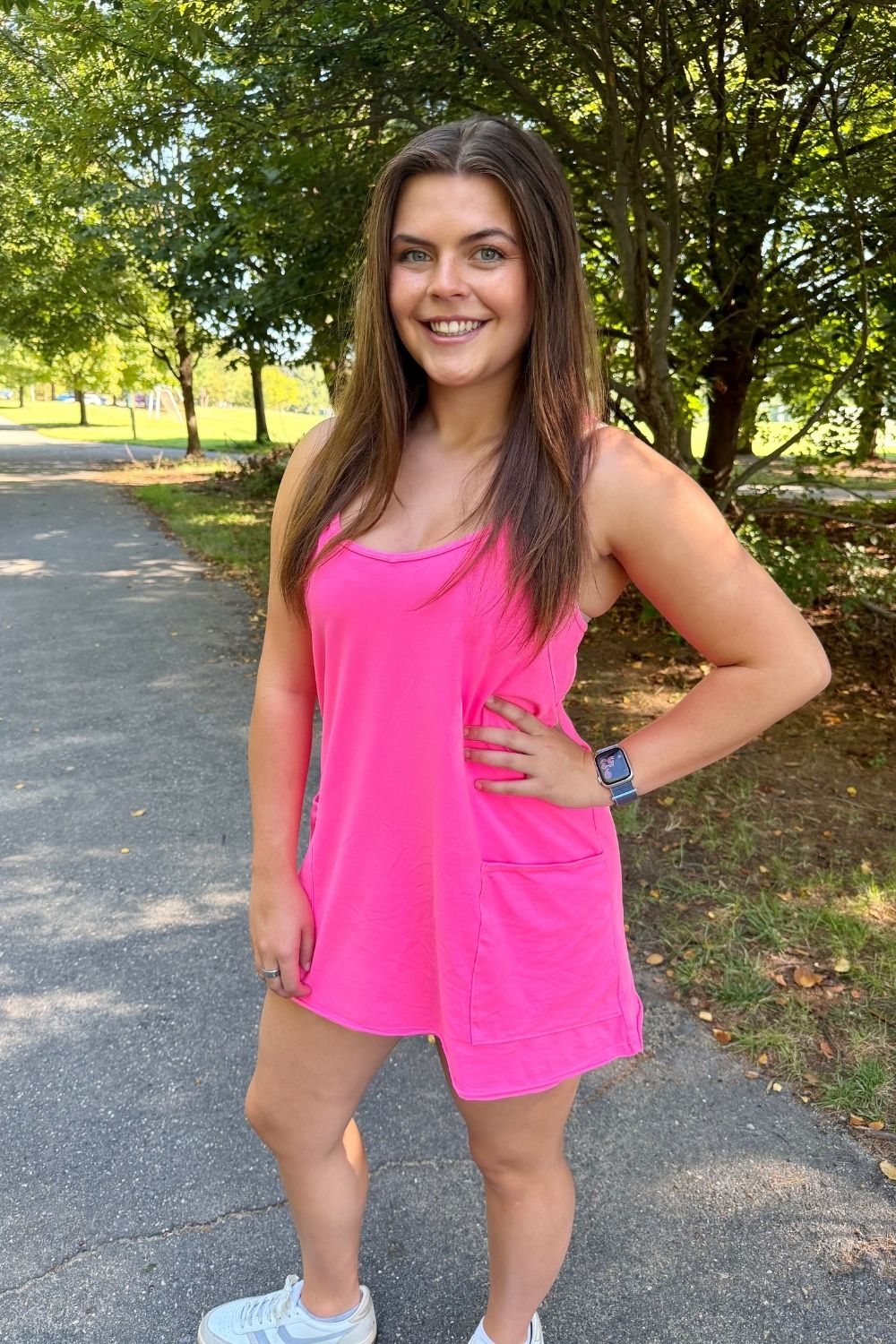A woman posing outdoors on a paved path in a park, wearing a bright pink sleeveless tankini-style mini dress with a front pocket.