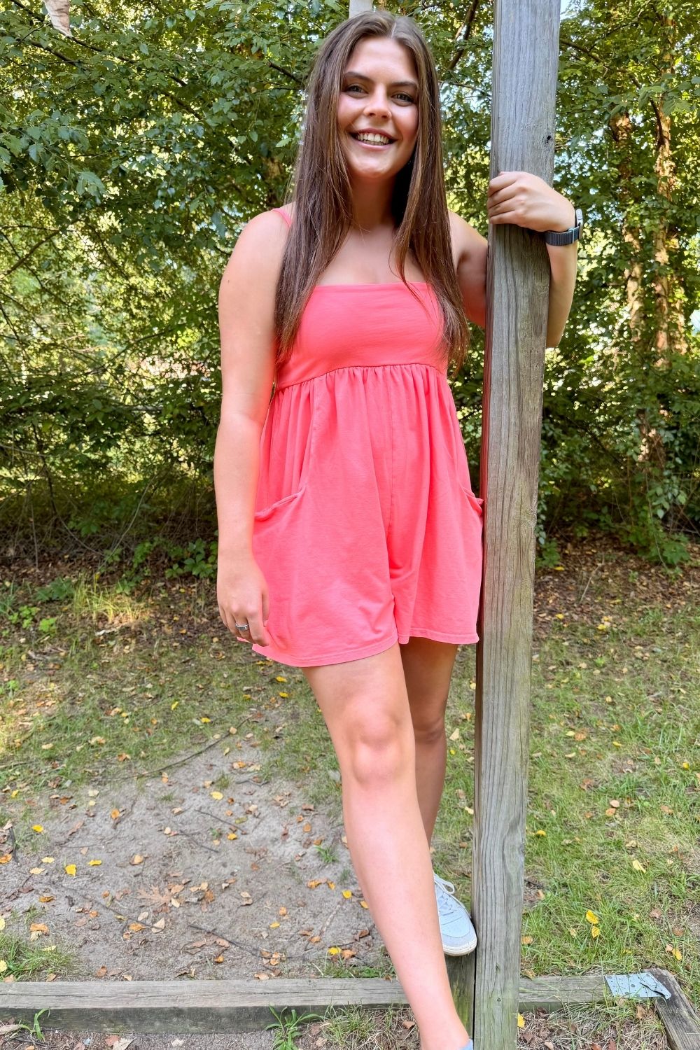 A woman wearing a coral pink sleeveless romper stands outdoors leaning against a wooden post, with trees and grass in the background.