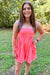 A woman wearing a bright coral pink strapless romper with side pockets, standing outdoors on a grassy lawn with trees in the background.