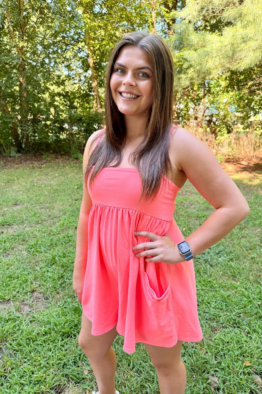 A woman wearing a bright coral pink sleeveless mini dress with side pockets, standing outdoors on a grassy lawn with trees in the background.