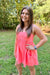 A woman wearing a bright coral pink sleeveless mini dress with side pockets, standing outdoors on a grassy lawn with trees in the background.