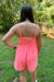 A woman viewed from behind wearing a bright coral pink romper with thin shoulder straps and a gathered waist, standing outdoors on grass with trees in the background.