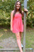 A woman wearing a coral pink sleeveless romper stands outdoors leaning against a wooden post, with trees and grass in the background.