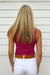 A woman viewed from behind wearing a fitted sleeveless magenta top, white pants, and a brown belt, standing against a white siding wall.