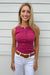 A smiling blonde woman wearing a sleeveless henley top in magenta pink, white pants, and a brown belt, standing against a white siding wall.