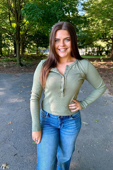A woman with long brown hair stands outdoors on a paved path wearing a ribbed long-sleeve henley top in sage green and blue jeans, with trees in the background.