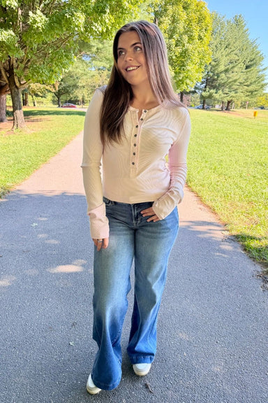 A woman stands on an outdoor paved path wearing a light beige long-sleeve Henley top and wide-leg blue jeans, with trees and grass in the background.