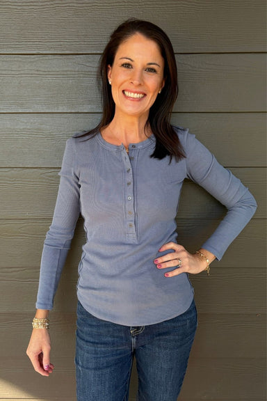 A smiling woman with dark hair stands against a gray wood-paneled wall, wearing a dusty blue long-sleeve henley top and blue jeans.