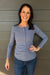 A smiling woman with dark hair stands against a gray wood-paneled wall, wearing a dusty blue long-sleeve henley top and blue jeans.
