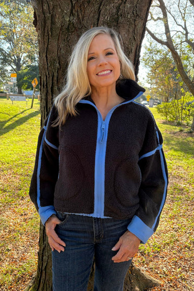 A woman with blonde hair stands outdoors in front of a tree wearing a dark brown fleece zip-up jacket with light blue trim and matching blue cuffs and collar, paired with dark blue jeans.