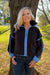 A woman with blonde hair stands outdoors in front of a tree wearing a dark brown fleece zip-up jacket with light blue trim and matching blue cuffs and collar, paired with dark blue jeans.