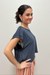 A woman modeling a short-sleeve heathered slate blue t-shirt with a side cutout, paired with a striped skirt, standing against a plain light gray wall.