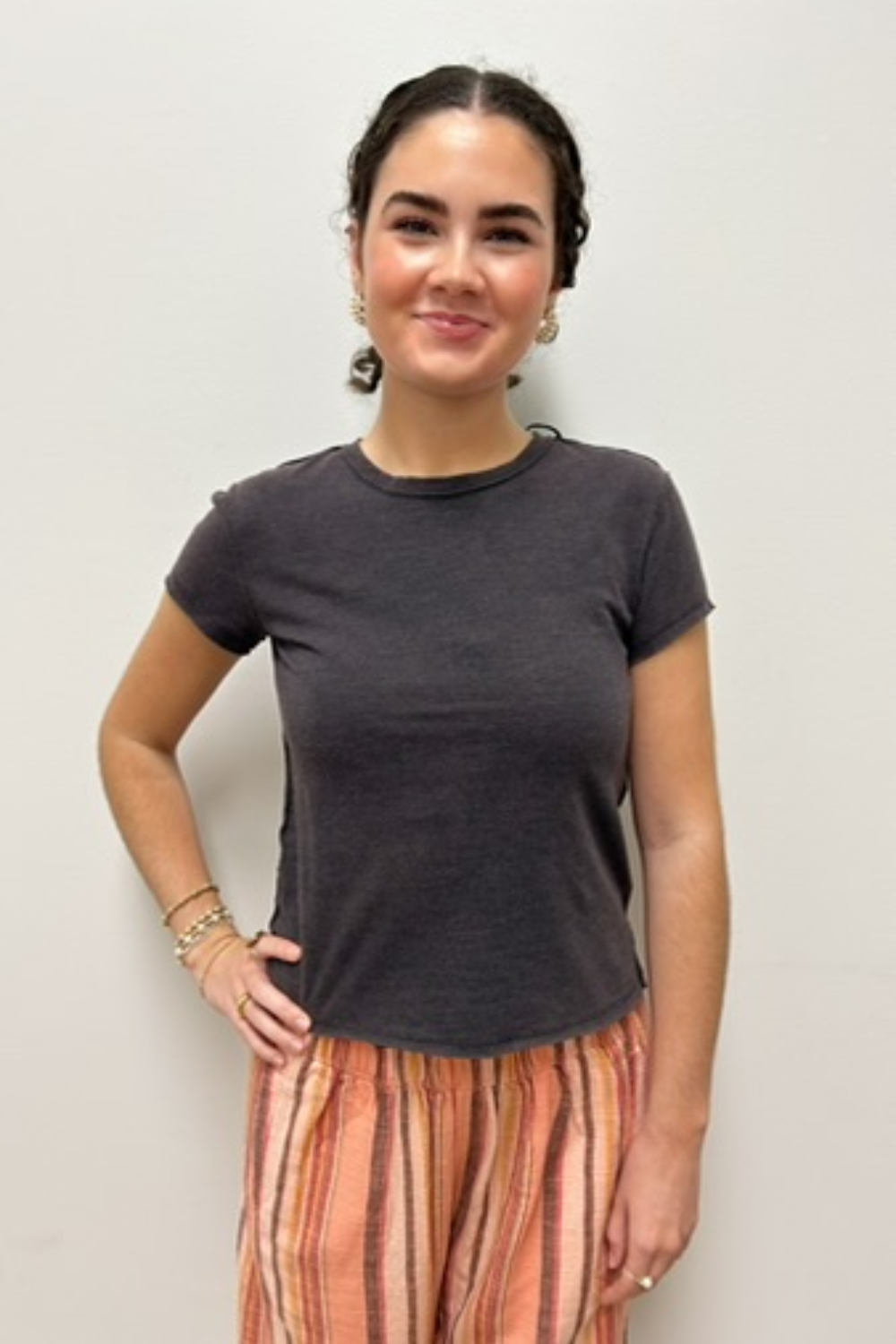 A woman modeling a dark charcoal gray short-sleeve crewneck t-shirt paired with orange and beige striped pants, standing against a plain light gray wall.