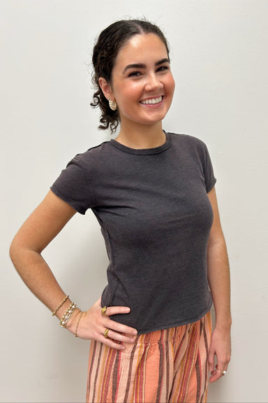 A woman smiling and posing in a dark charcoal gray short-sleeve crewneck t-shirt and a multi-colored striped skirt against a plain light gray background.