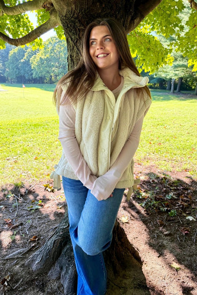 A woman wearing a cream fleece vest over a light pink long-sleeve top and blue jeans, leaning against a tree in a sunny park.