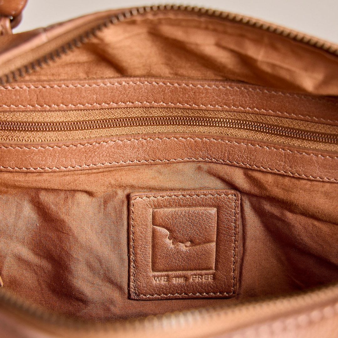 Close-up of the interior of a brown leather bag, showing a zippered compartment, stitched leather trim, and a square embossed brand patch that reads 'WE DON'T FEEL FREE'.