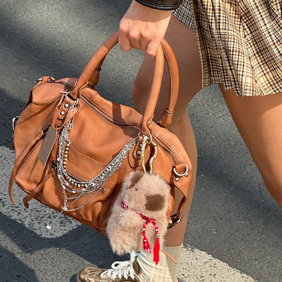 A person carrying a tan leather shoulder bag outdoors on asphalt. The bag has silver chain and bead embellishments, a fluffy beige dog-shaped charm with a red bandana, and is styled with a plaid skirt and sneakers.