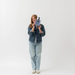 A woman stands against a plain white studio background wearing a blue denim jacket, light blue jeans, and white sneakers, while holding up a small striped accessory with a white feather.