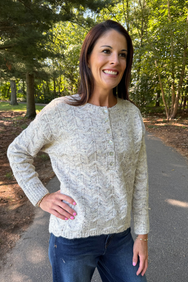 A woman wearing a cream cable-knit button-up sweater and blue jeans, standing on an outdoor paved path surrounded by trees.