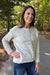 A woman wearing a cream cable-knit button-up sweater and blue jeans, standing on an outdoor paved path surrounded by trees.