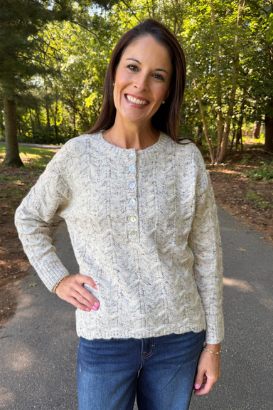 A woman smiling outdoors on a paved path wearing a cream cable-knit long-sleeve henley sweater with blue jeans.