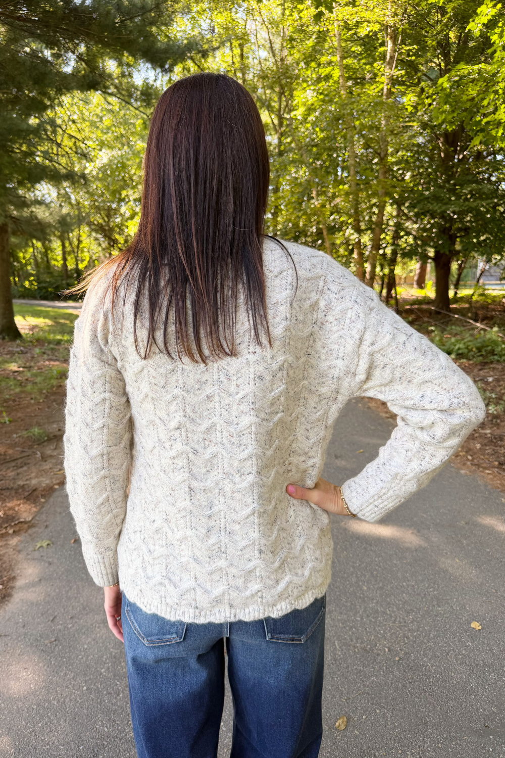 A woman viewed from behind wearing a cream cable-knit sweater and blue jeans, standing on a paved path in a wooded outdoor setting.
