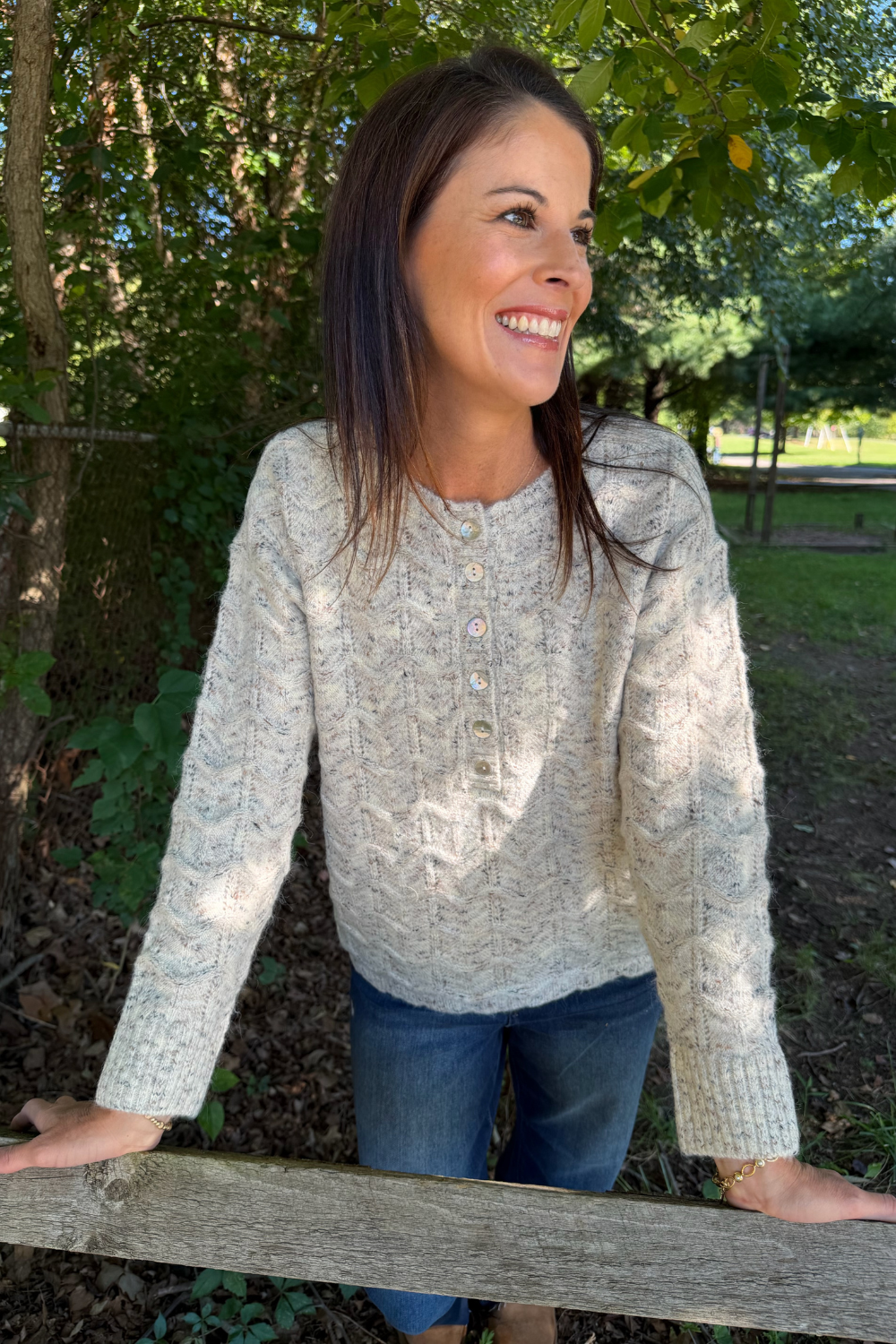 A woman smiling outdoors in a light beige cable-knit henley sweater with blue jeans, leaning on a wooden fence in a wooded setting.