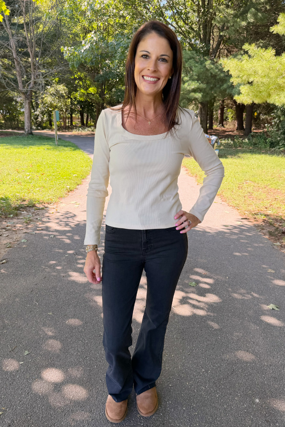 A woman stands on a paved path in a park wearing a cream ribbed long-sleeve scoop-neck top, dark charcoal pants, and tan boots.
