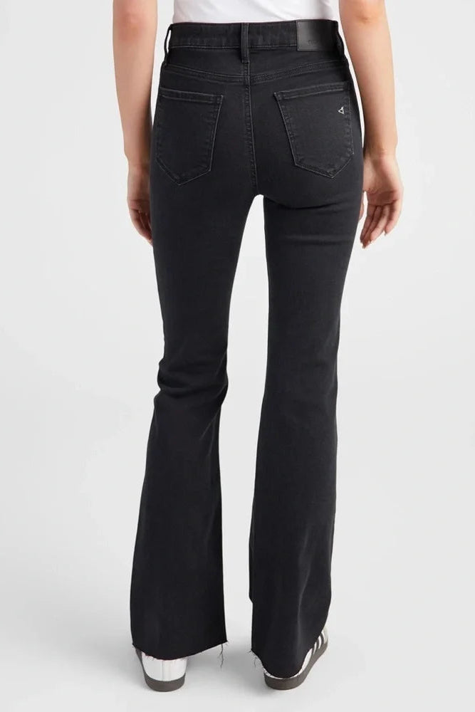 Rear view of a person wearing black flared jeans with a white top and white sneakers, photographed against a light gray studio background.
