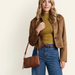 A woman modeling a cropped brown suede jacket over an olive green mock neck top, paired with blue jeans and a brown leather crossbody bag, against a plain beige background.