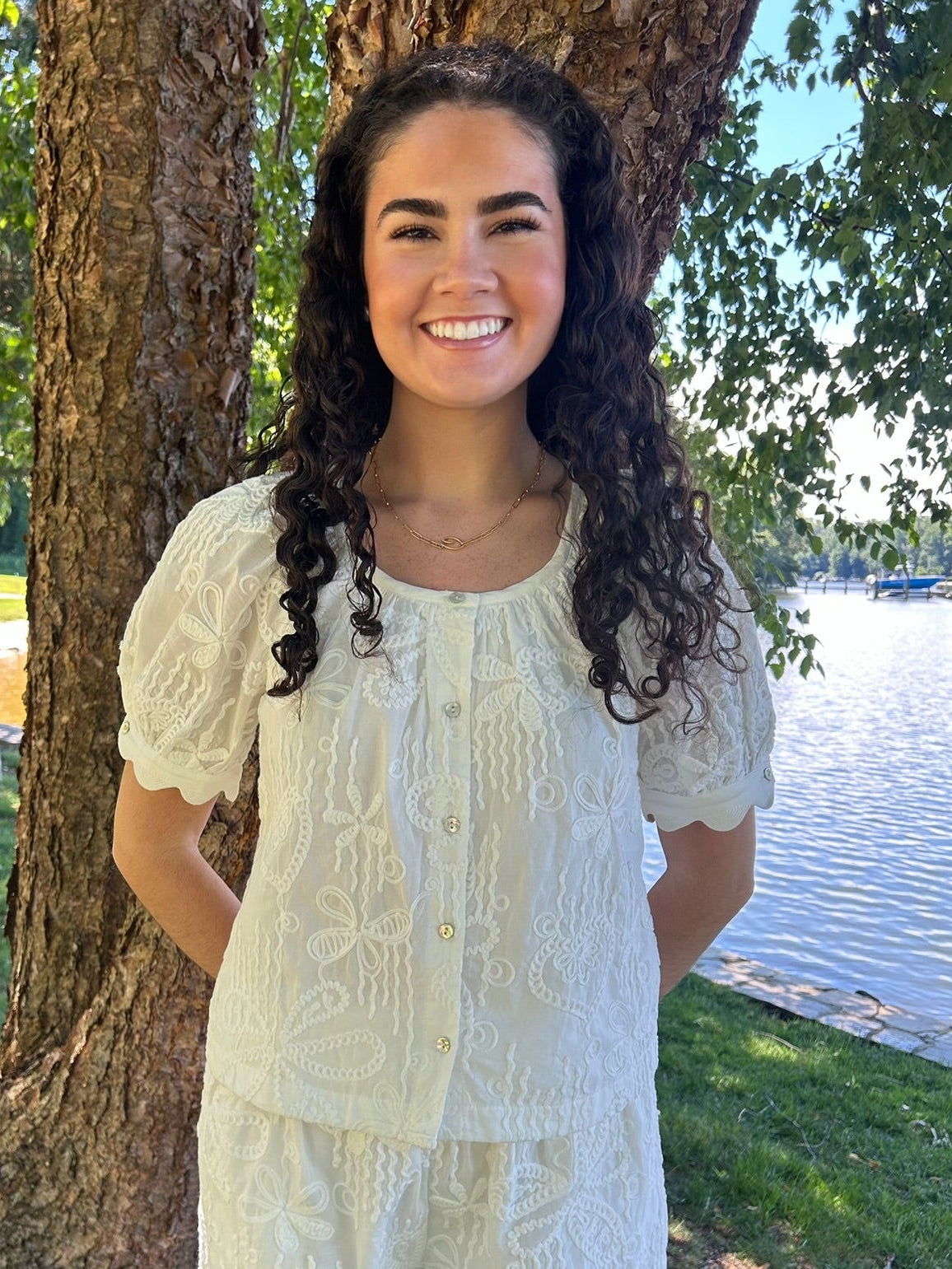 Woman in a light-colored outfit standing by a tree by a body of water