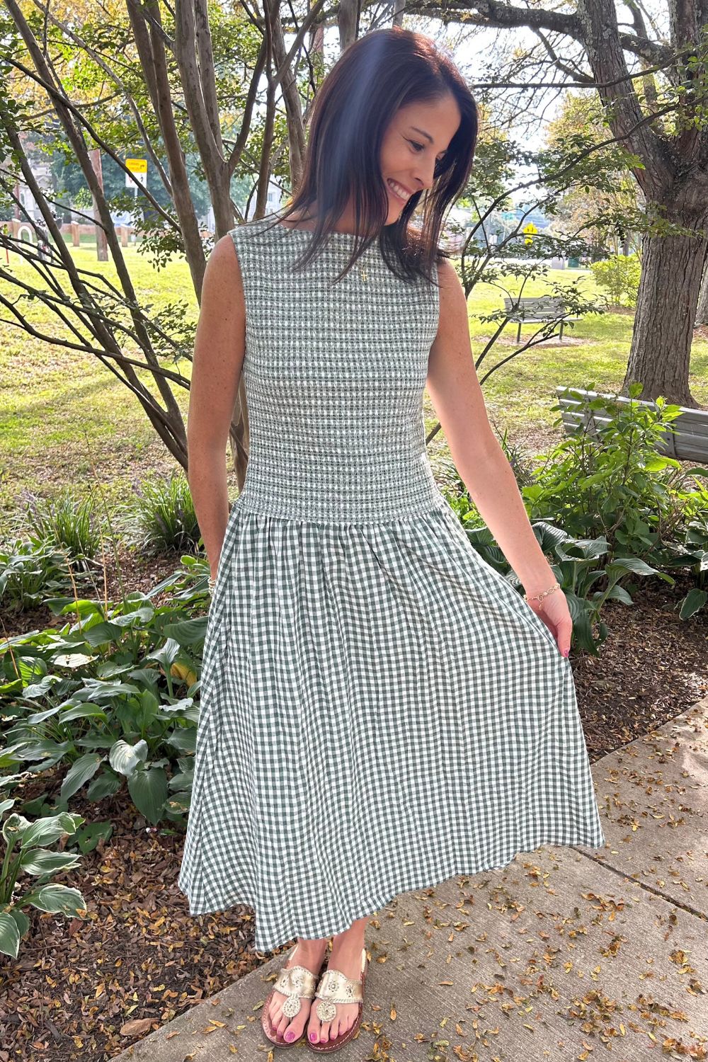 A woman wearing a sleeveless green and white gingham midi dress stands outdoors on a paved path surrounded by trees and greenery, holding out one side of the skirt to show its flow.