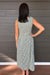 A woman viewed from behind wearing a sleeveless green and white gingham midi dress, standing against a dark gray wood-paneled wall.