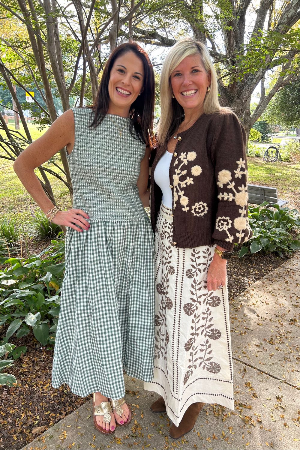 Two women standing outdoors on a paved path surrounded by trees and greenery, wearing coordinated outfits. The woman on the left wears a sleeveless green-and-white gingham dress, and the woman on the right wears a brown floral cardigan over a white top with a long cream skirt featuring brown floral embroidery.