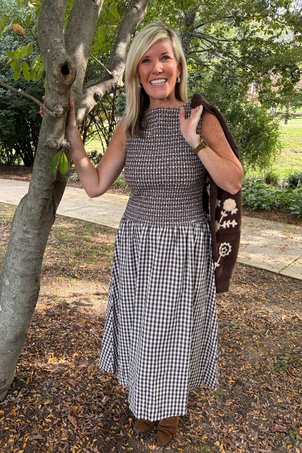 A woman stands outdoors next to a tree wearing a sleeveless smocked top and a matching gingham midi skirt, with a brown patterned shawl draped over her shoulder.