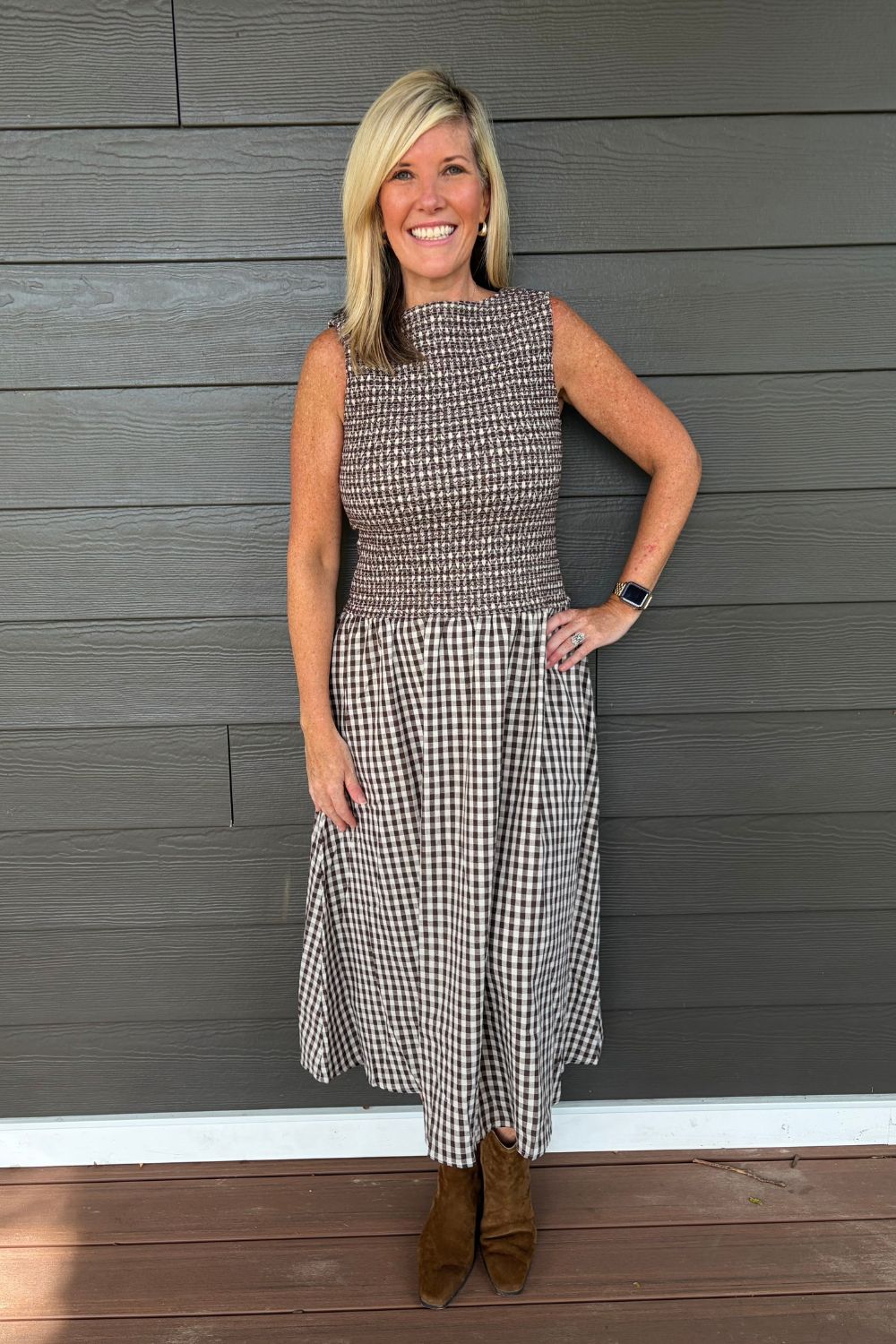 A woman wearing a sleeveless green and white gingham midi dress stands outdoors on a paved path surrounded by trees and greenery, holding out one side of the skirt to show its flow.