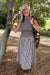 A woman stands outdoors next to a tree wearing a sleeveless smocked top and a matching gingham midi skirt, with a brown patterned shawl draped over her shoulder.