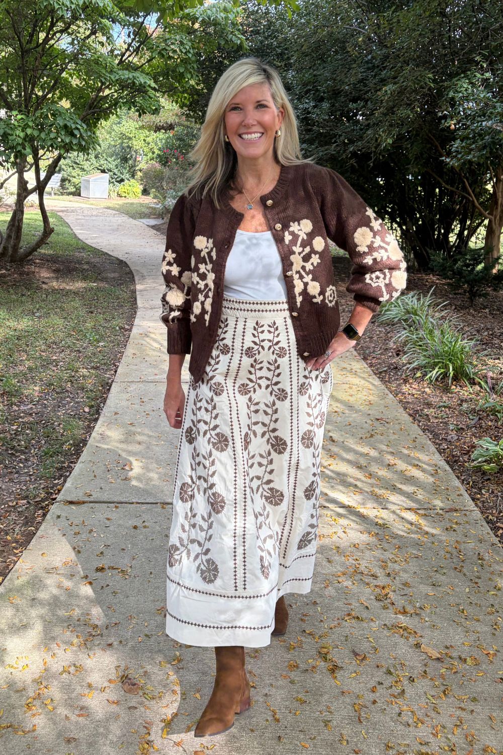 A woman stands on a paved path outdoors wearing a brown embroidered cardigan over a white tank top and a matching white maxi skirt with brown floral embroidery, paired with tan ankle boots.