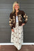 A person wearing a cream maxi skirt with brown floral embroidery and a matching brown cardigan, standing against a gray wood-paneled wall.