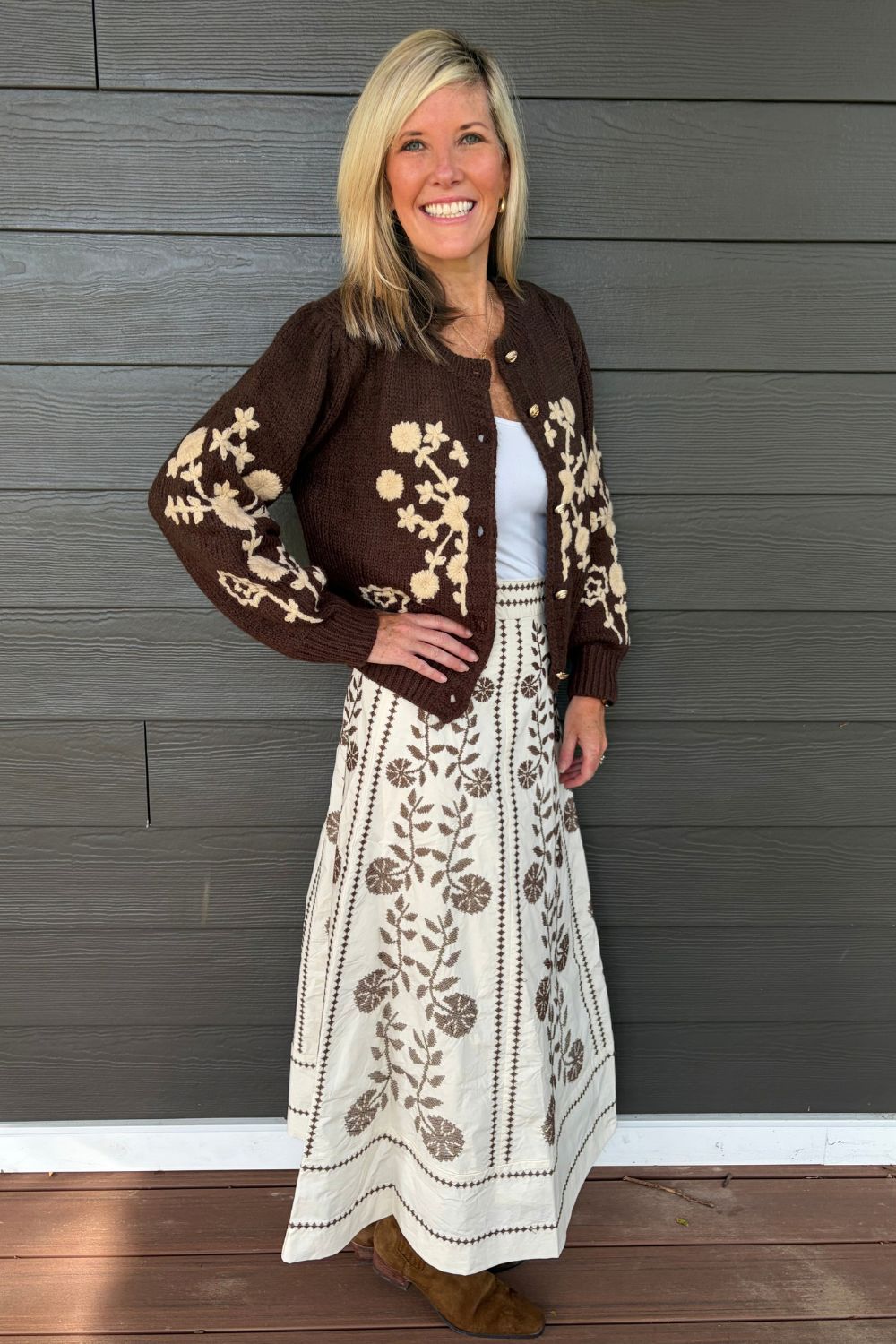 A woman modeling a brown and cream floral knit cardigan over a white top and a matching cream skirt with brown floral embroidery, standing against a dark gray wood-paneled wall.