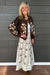 A woman modeling a brown and cream floral knit cardigan over a white top and a matching cream skirt with brown floral embroidery, standing against a dark gray wood-paneled wall.