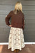 A woman viewed from behind wearing a brown cropped knit sweater with beige patchwork details on the sleeves and a cream-colored midi skirt with a brown floral and geometric leaf pattern. She is standing against a gray wood-paneled wall on a wooden deck.