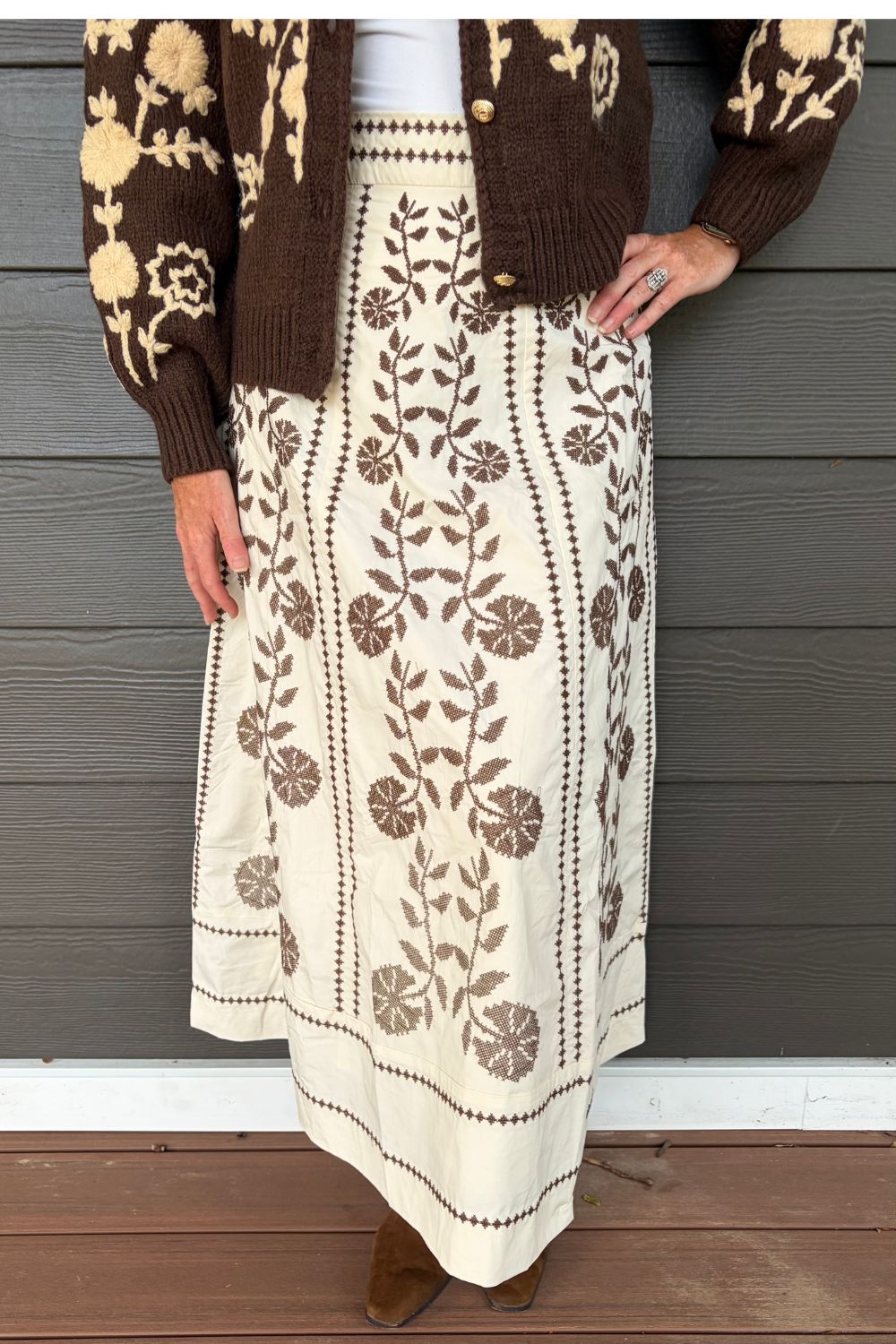 A person wearing a cream maxi skirt with brown floral embroidery and a matching brown cardigan, standing against a gray wood-paneled wall.
