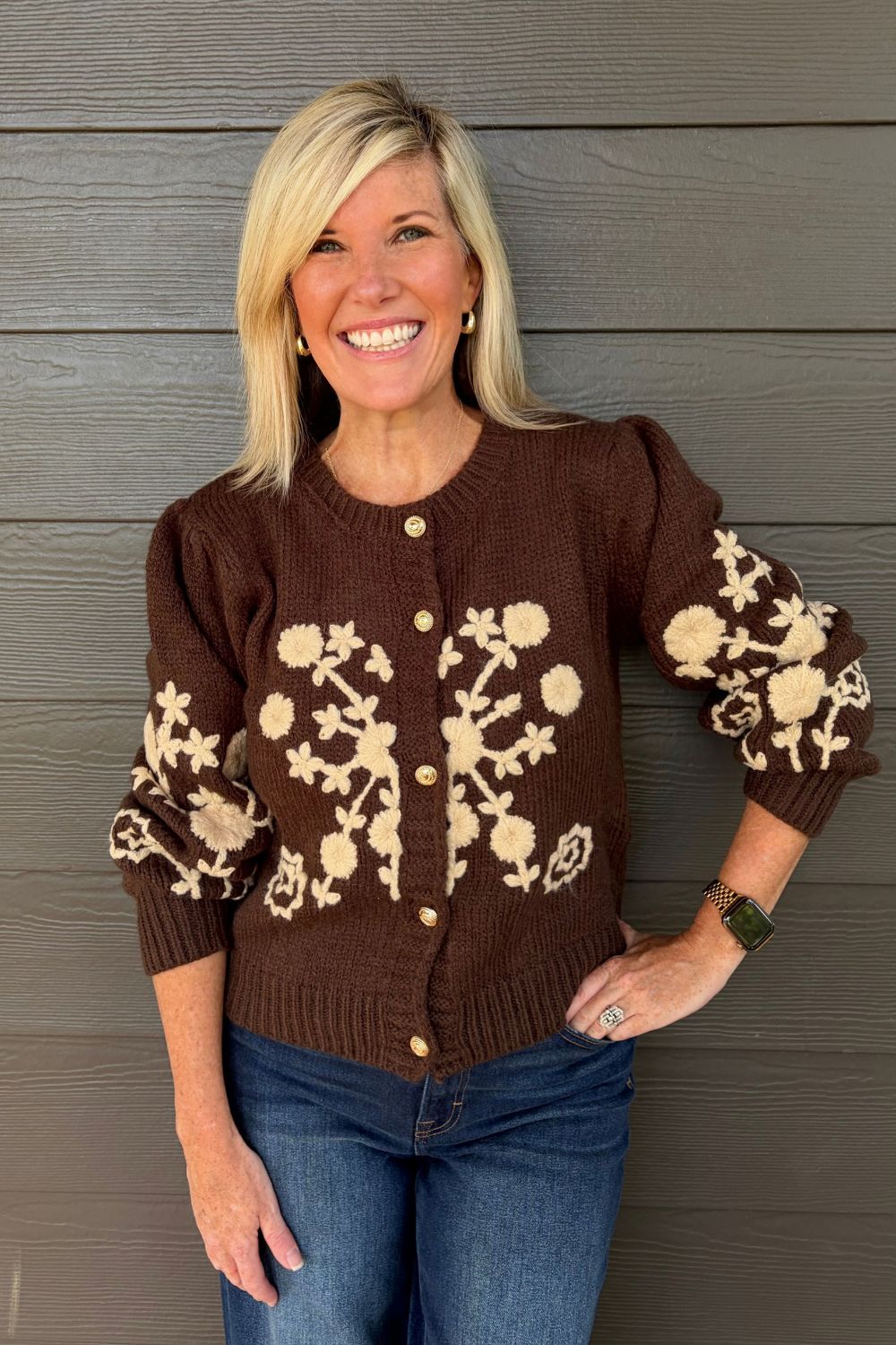 A smiling blonde woman wearing a brown knit cardigan with beige floral and leaf patterns on the front and sleeves, paired with blue jeans, standing against a dark gray wood-paneled wall.