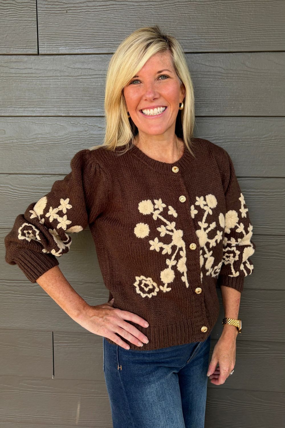 A smiling blonde woman wearing a brown knit cardigan with cream floral patterns and gold buttons, paired with blue jeans, standing against a gray wood-paneled wall.