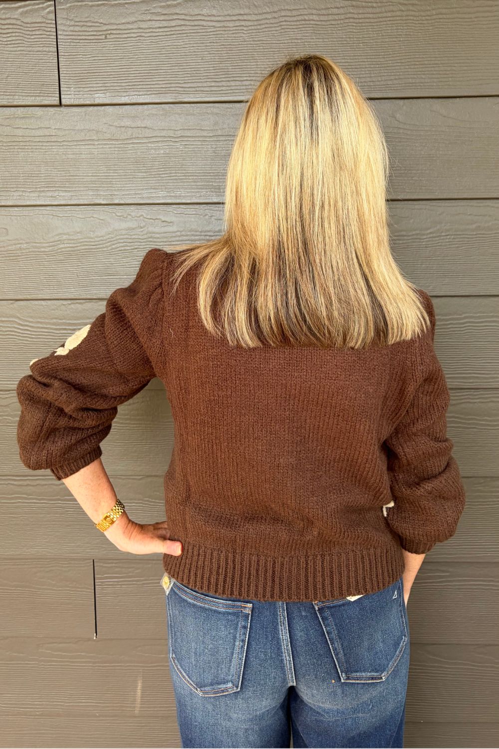A blonde woman wearing a brown knit cardigan, paired with blue jeans, standing against a gray wood-paneled wall.