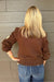 A blonde woman wearing a brown knit cardigan, paired with blue jeans, standing against a gray wood-paneled wall.