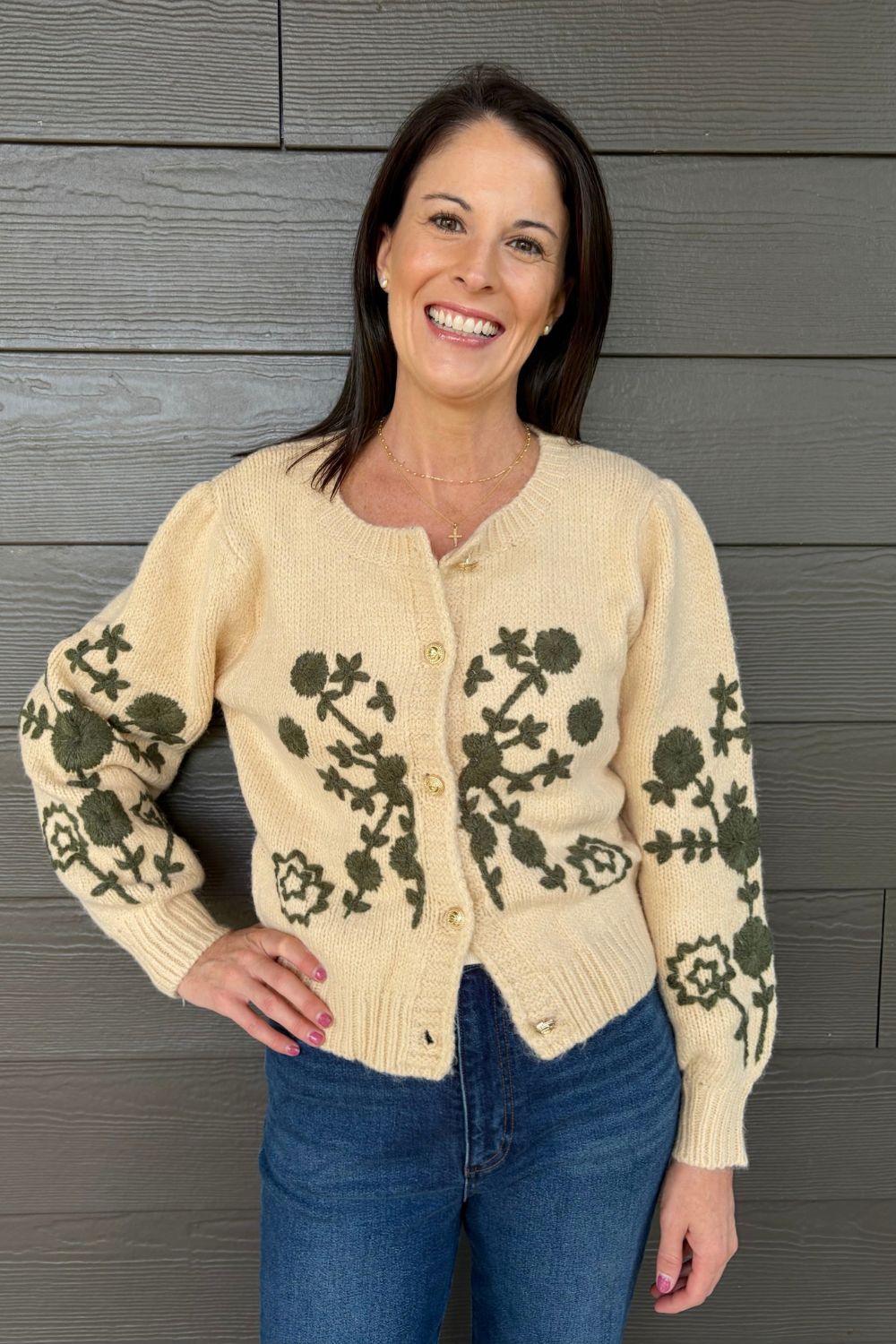 A smiling woman wearing a cream-colored knit cardigan with dark green floral and botanical patterns, paired with blue jeans, standing against a gray wood-paneled wall.
